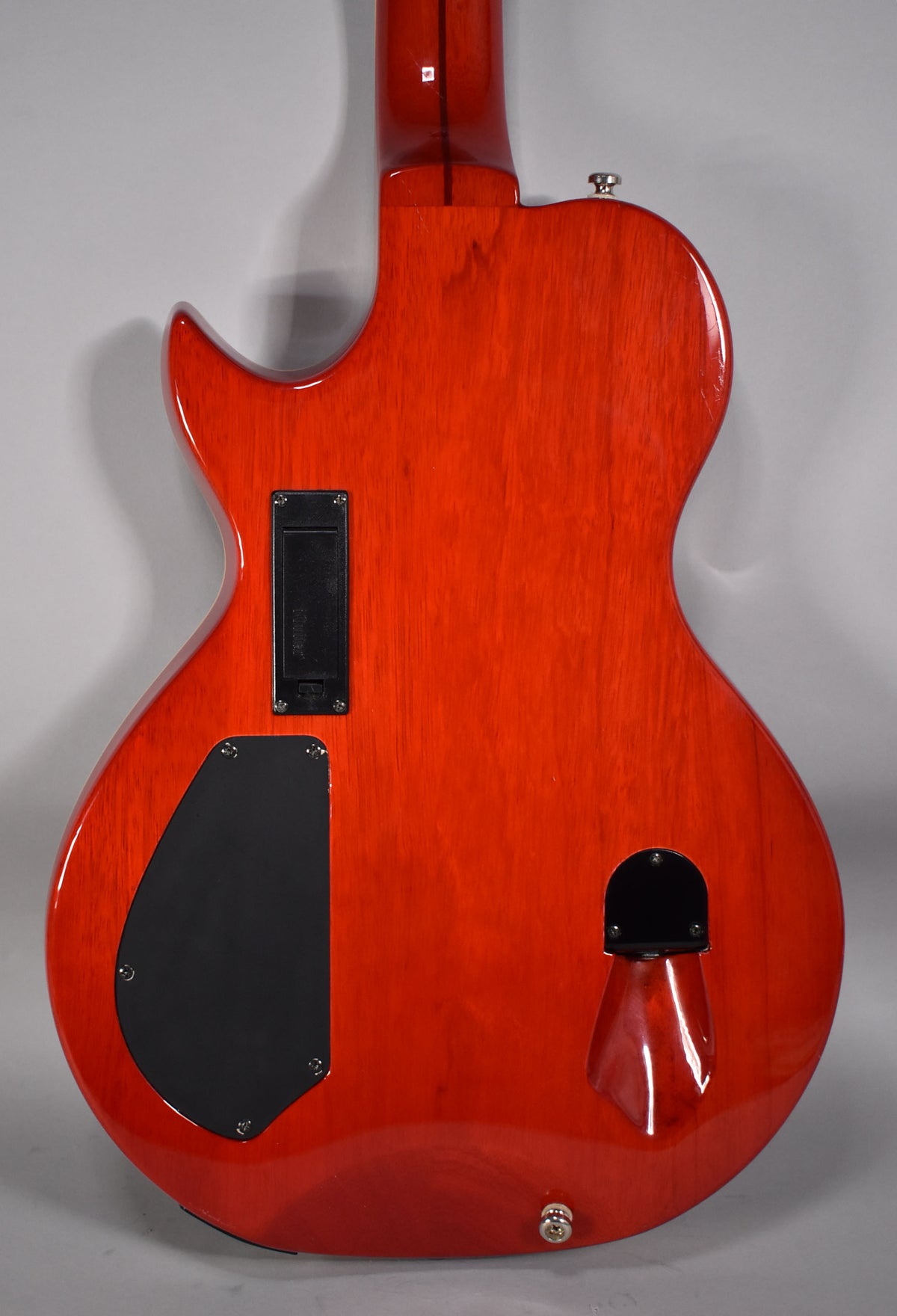 The back of a glossy cinnamon-red electric guitar, showing black control plates, a battery compartment, and a chrome strap button.