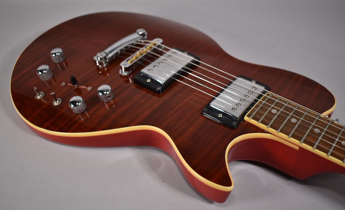 The cinnamon-finished electric guitar is angled from the upper left, showing its flamed top, chrome hardware, and cream binding.