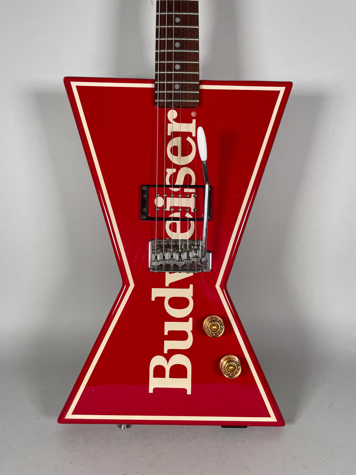 The red bowtie-shaped guitar body, with cream trim and "Budweiser" text, features a single humbucker, tremolo, and two gold knobs.