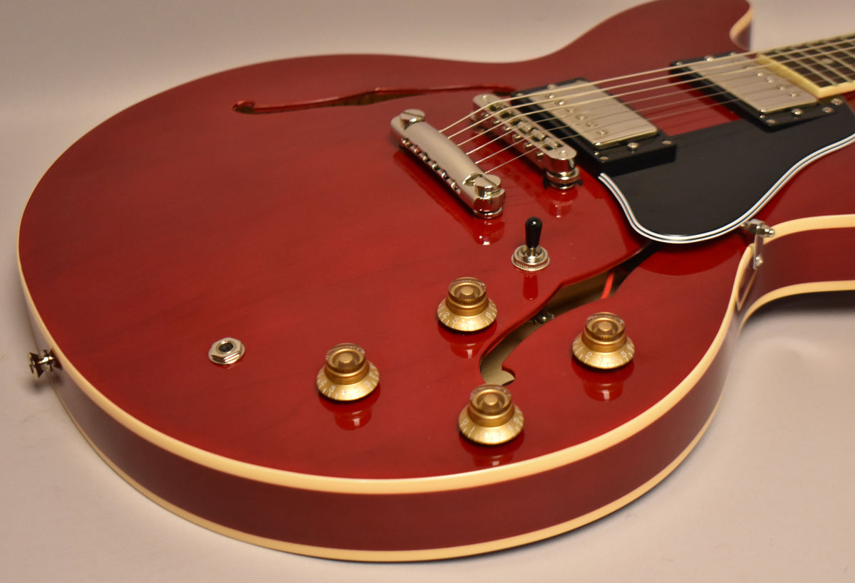 The cherry red semi-hollow body guitar, with cream binding, shows its lower bout, four gold control knobs, and a three-way toggle switch.
