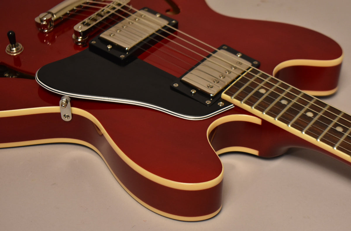 Close-up of the cherry red semi-hollow body with cream binding, black pickguard, two chrome humbuckers, and part of the fretboard.