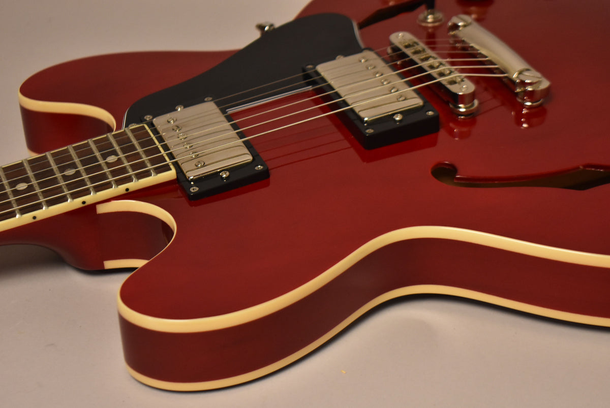 Close-up of a cherry red semi-hollow electric guitar, showing the lower bout, neck, two humbuckers, and a black pickguard.