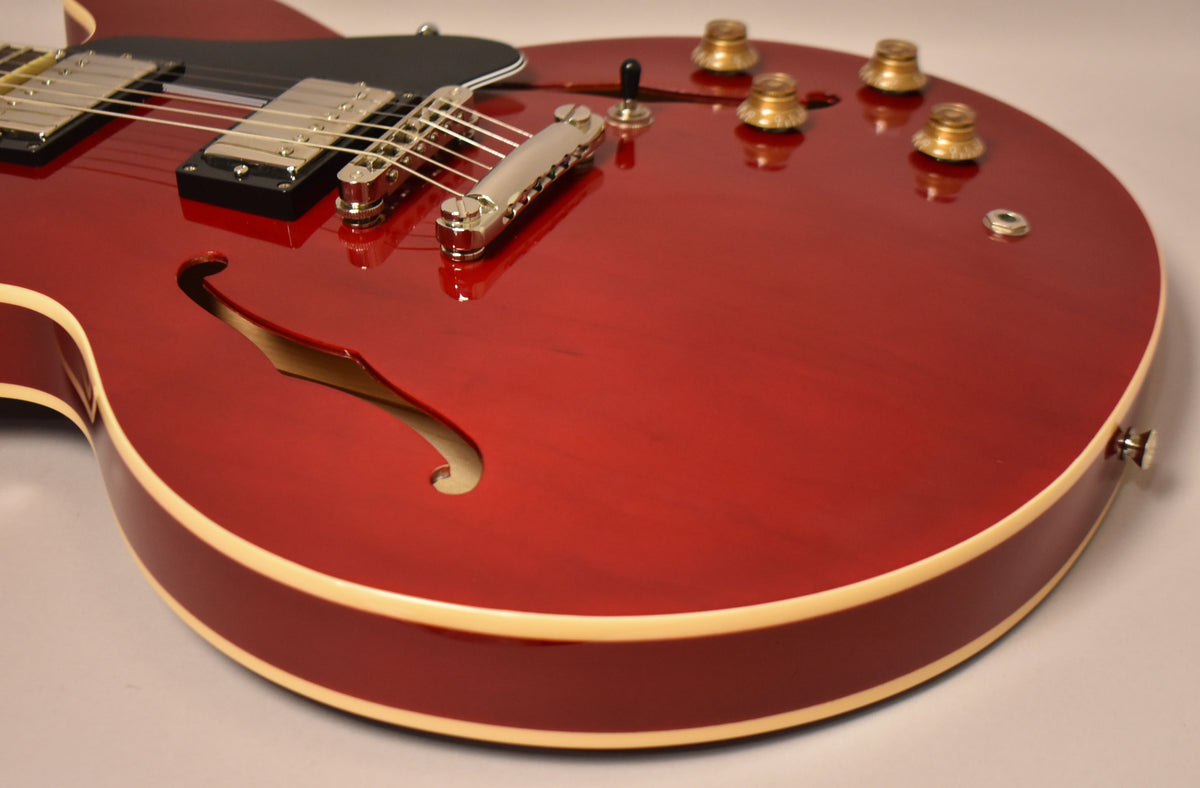 The cherry red hollow-body guitar, with cream binding and f-hole, shows a black pickguard, chrome hardware, and four gold knobs.