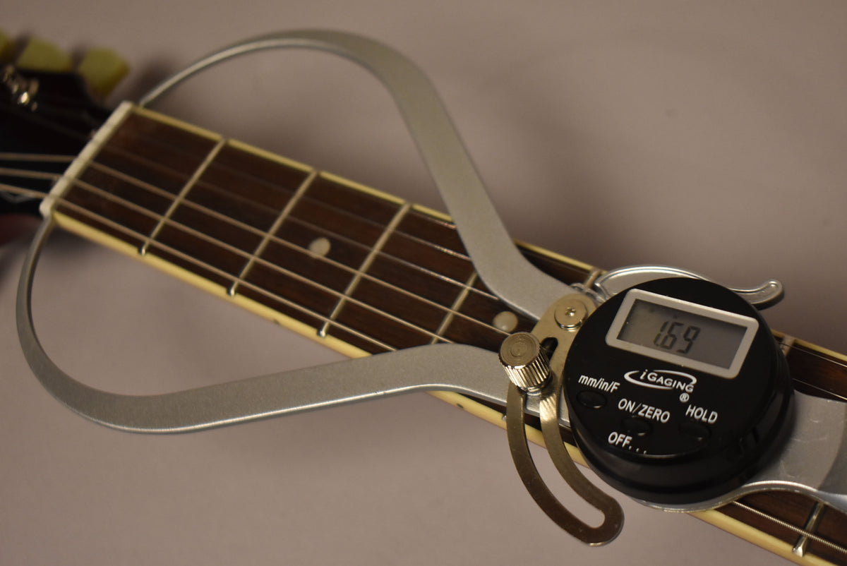 A digital caliper measures the guitar's nut width at 1.69 inches, resting on the dark fretboard with cream binding.
