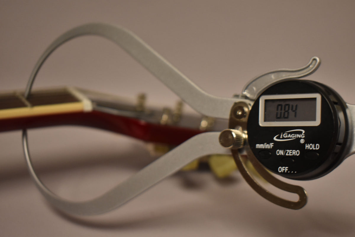A digital caliper measures the guitar's cherry red neck, displaying "0.84" on its screen, with tuning pegs visible in the background.