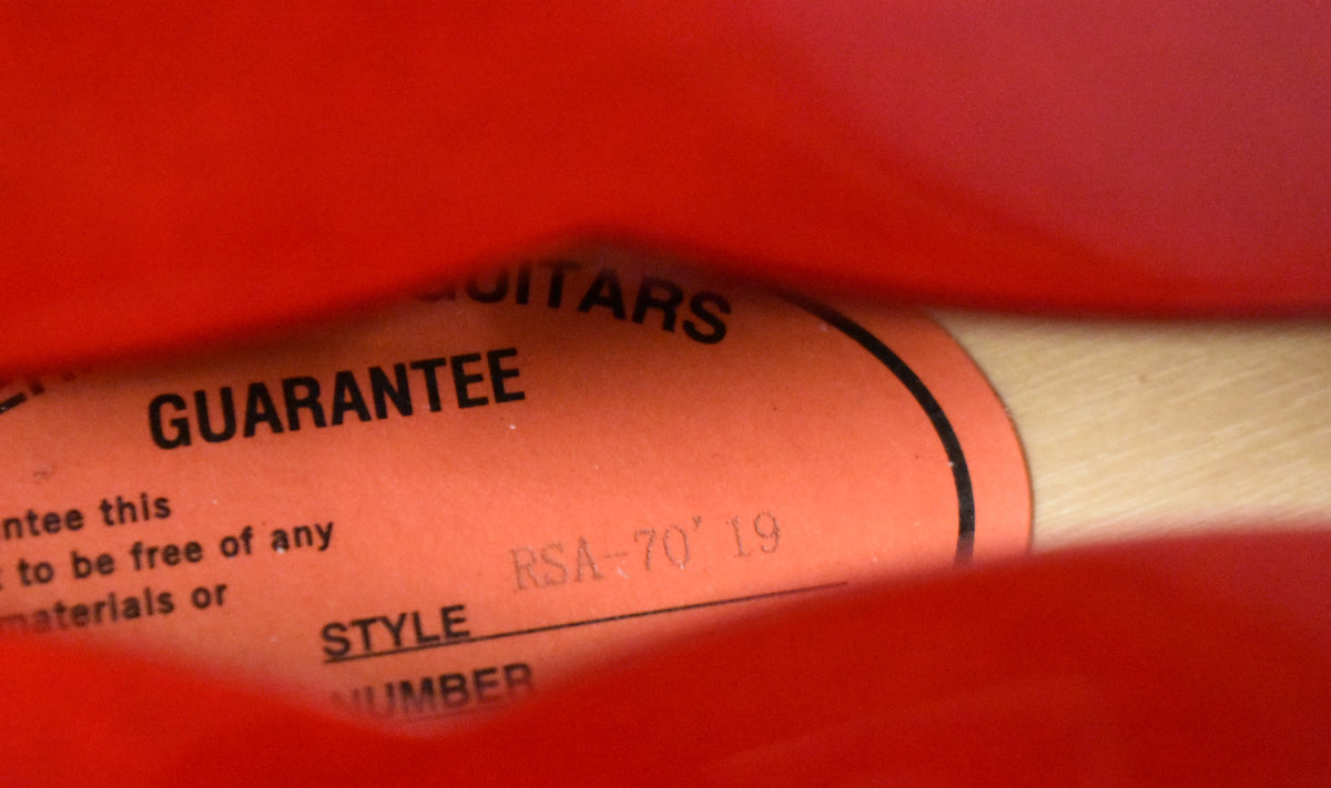 Close-up of a red paper guarantee card with "RSA-70 '19" handwritten, partially obscured by the red gig bag.