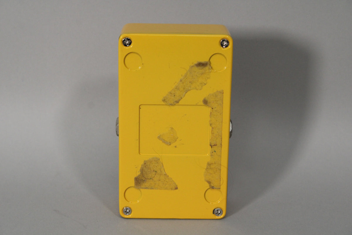 The bright yellow pedal's underside is visible, showing four silver screws, a recessed rectangle, and remnants of adhesive.