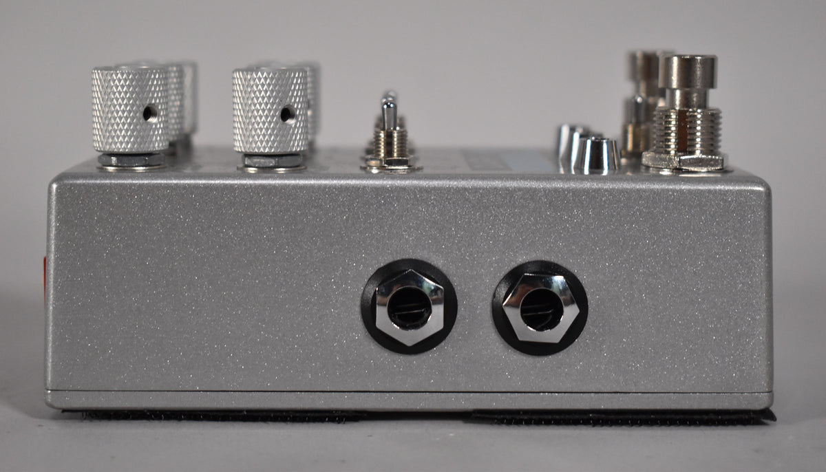 The silver glitter-finish pedal is viewed from the right side, showing two input jacks and several silver knobs and switches.