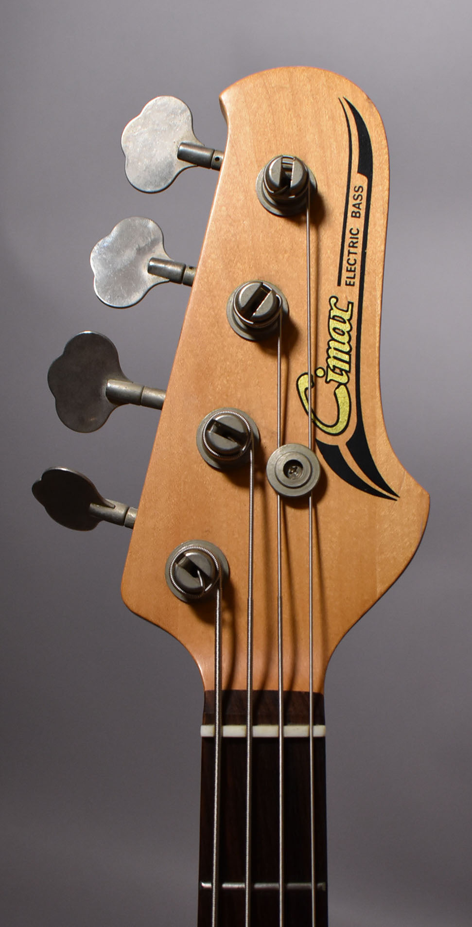 Close-up of the bass headstock, showing the light wood finish, four metal tuners, and the top of the dark fretboard.