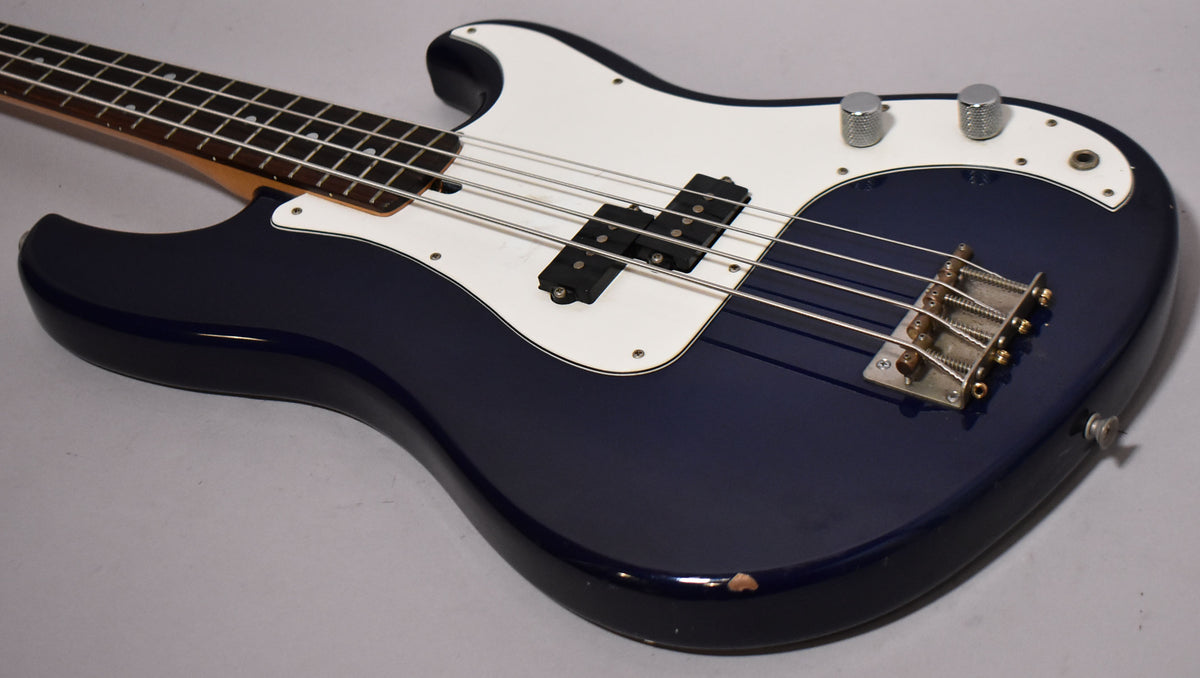Indigo blue bass, angled from the neck to the lower bout, showing a white pickguard, P/J pickups, and a chip on the lower edge.