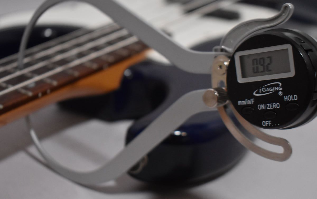 A digital caliper measures the string height at the 12th fret of the indigo blue bass, showing 0.92mm.