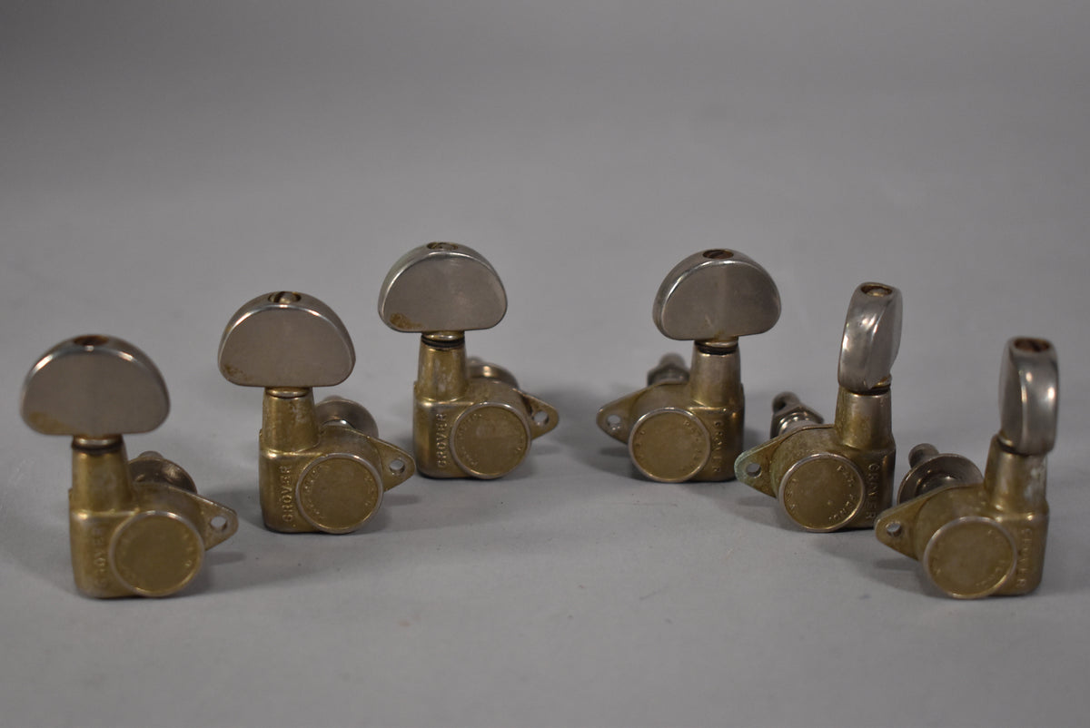 Six gold-plated tuners with tarnished, brushed metal buttons are arranged on a light gray surface, showing visible wear and patina.