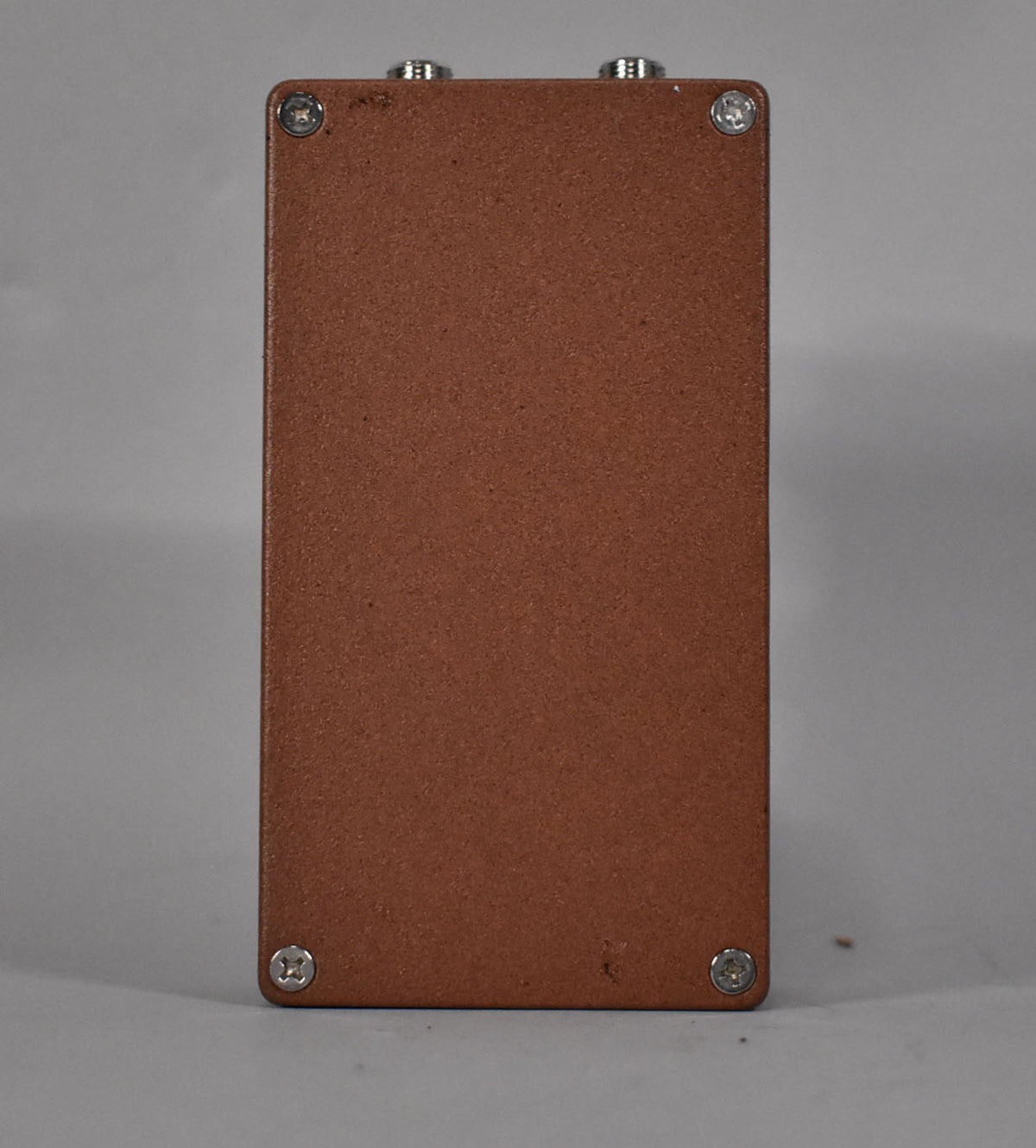 The brown pedal is viewed from the top, showing its textured finish, four silver screws, and two silver jacks at the far end.