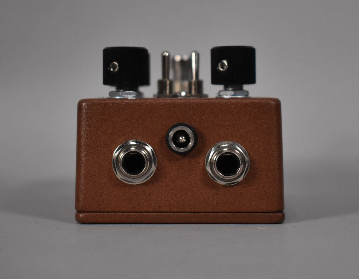 The brown pedal is viewed from the front, showing two input jacks, a power input, and two black control knobs on top.