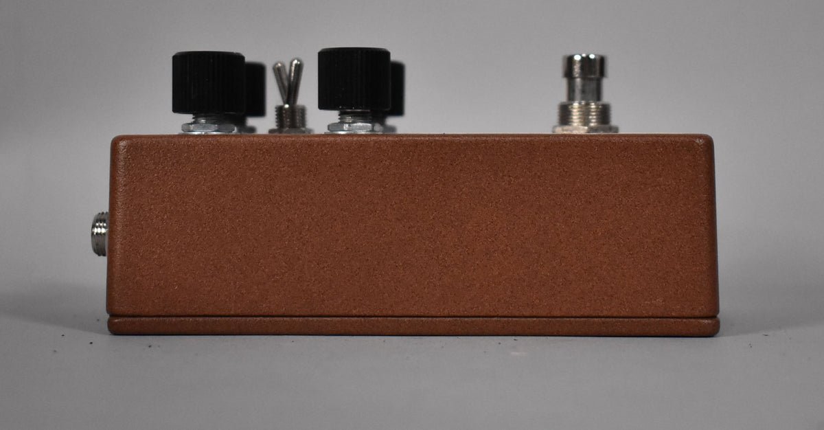 Side view of the brown textured pedal, showing two black knobs, a silver toggle switch, and a silver input jack.
