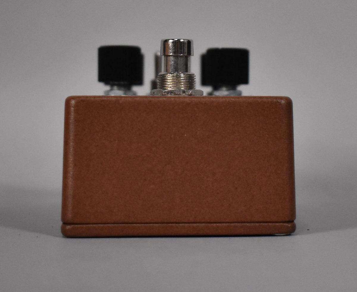 The brown pedal is viewed from the back, showing a textured finish and three black knobs and a silver jack on top.