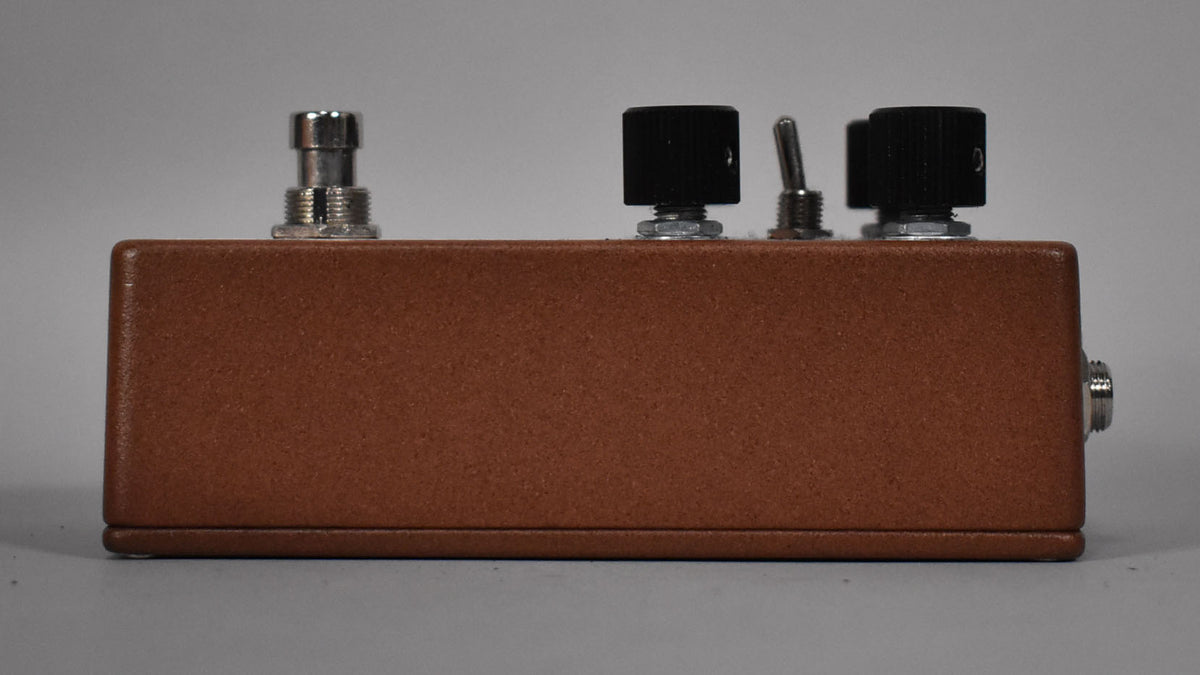 The brown pedal is viewed from the side, showing a silver footswitch, two black knobs, a silver toggle switch, and an input jack.