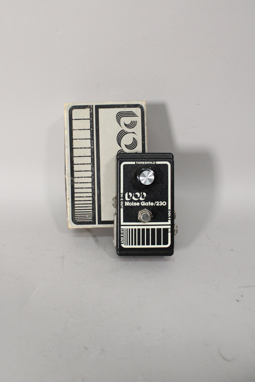Black pedal with a single silver knob and footswitch, showing some scuffs, next to its white box with black graphics.