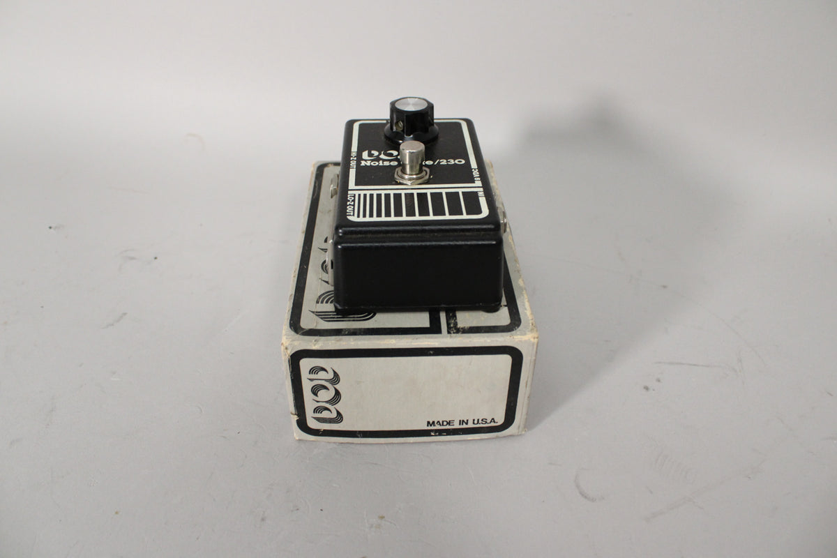 A black pedal with a single silver knob and footswitch sits on its worn, white and grey cardboard box, viewed from the front right.