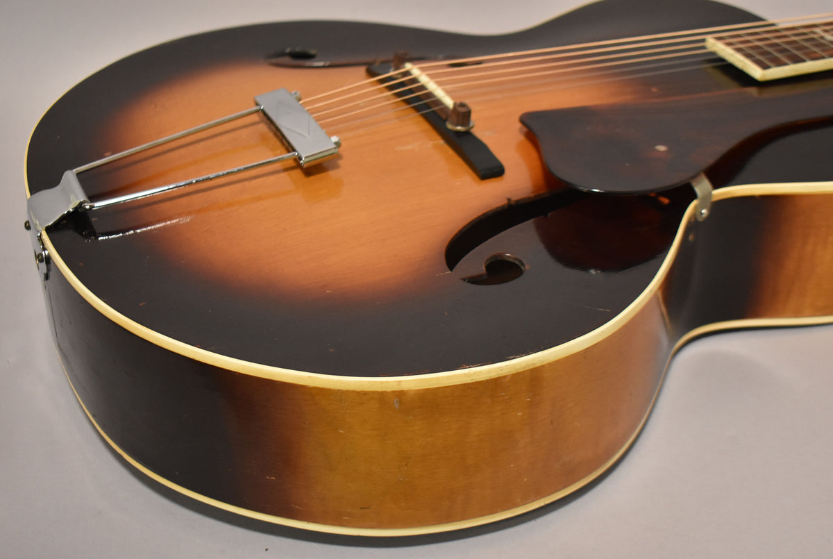 The archtop acoustic guitar's sunburst finish shows wear and checking, with a chrome trapeze tailpiece and dark pickguard.