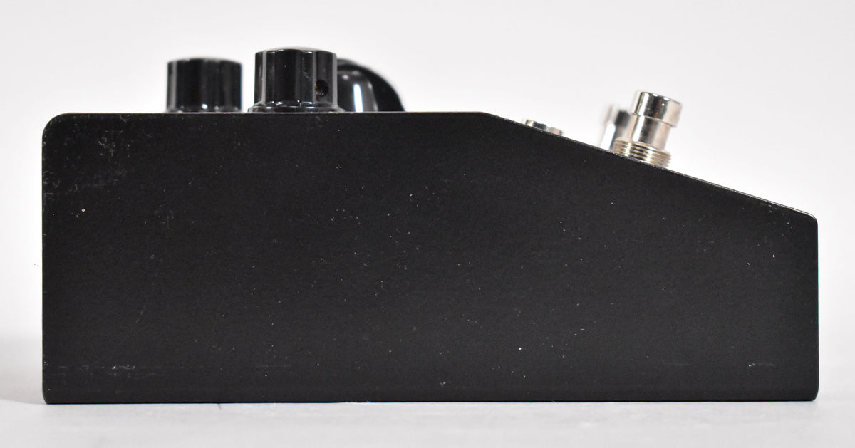 The black textured pedal is viewed from the right side, showing two black control knobs and a silver input jack on the top edge.