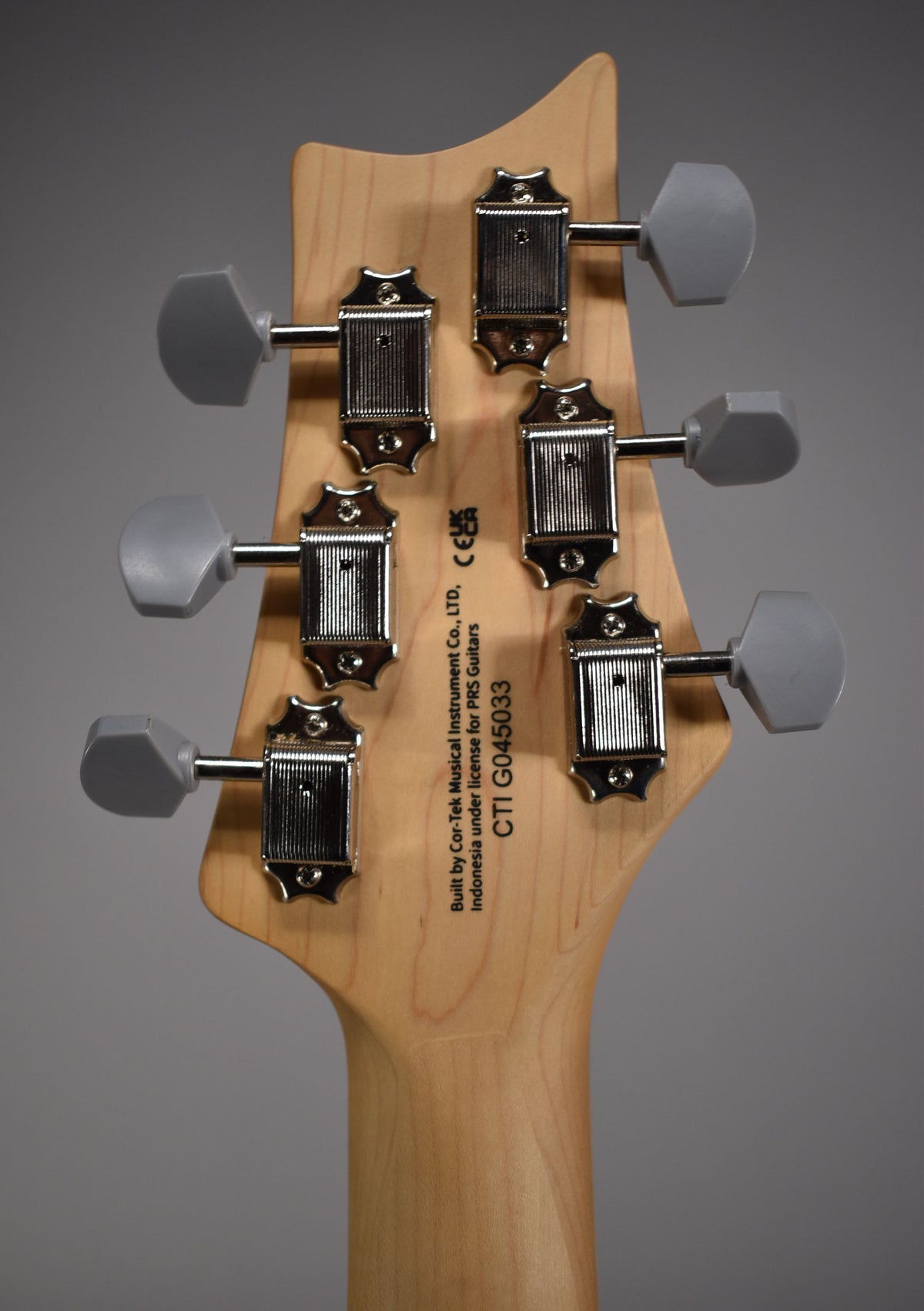 The back of a light wood headstock with six chrome tuners, each with a gray oval button, and visible serial number.