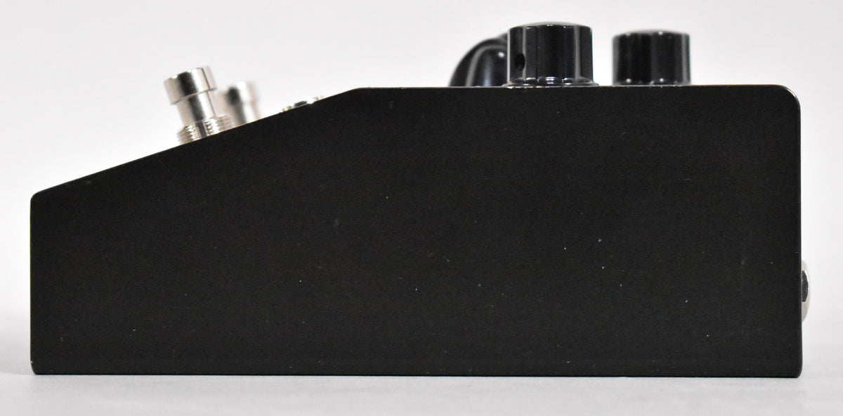 The black pedal is viewed from the right side, showing two silver input jacks and two black control knobs on the top edge.