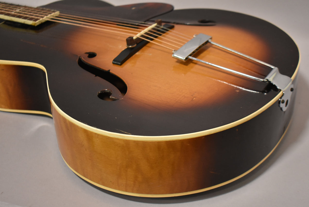 The archtop acoustic guitar's sunburst body, f-holes, and chrome trapeze tailpiece are visible, with visible wear and checking on the finish.