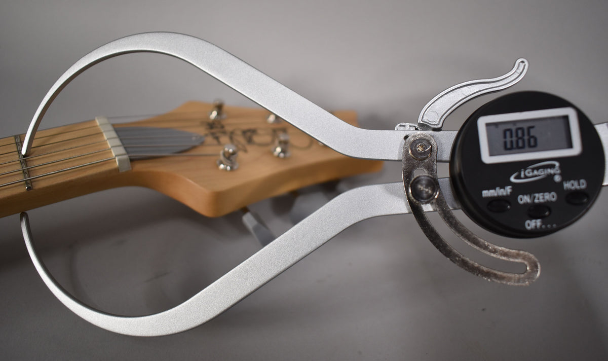 A digital caliper measures the guitar's nut width, showing "0.86" on its display, with the headstock and tuners blurred behind.