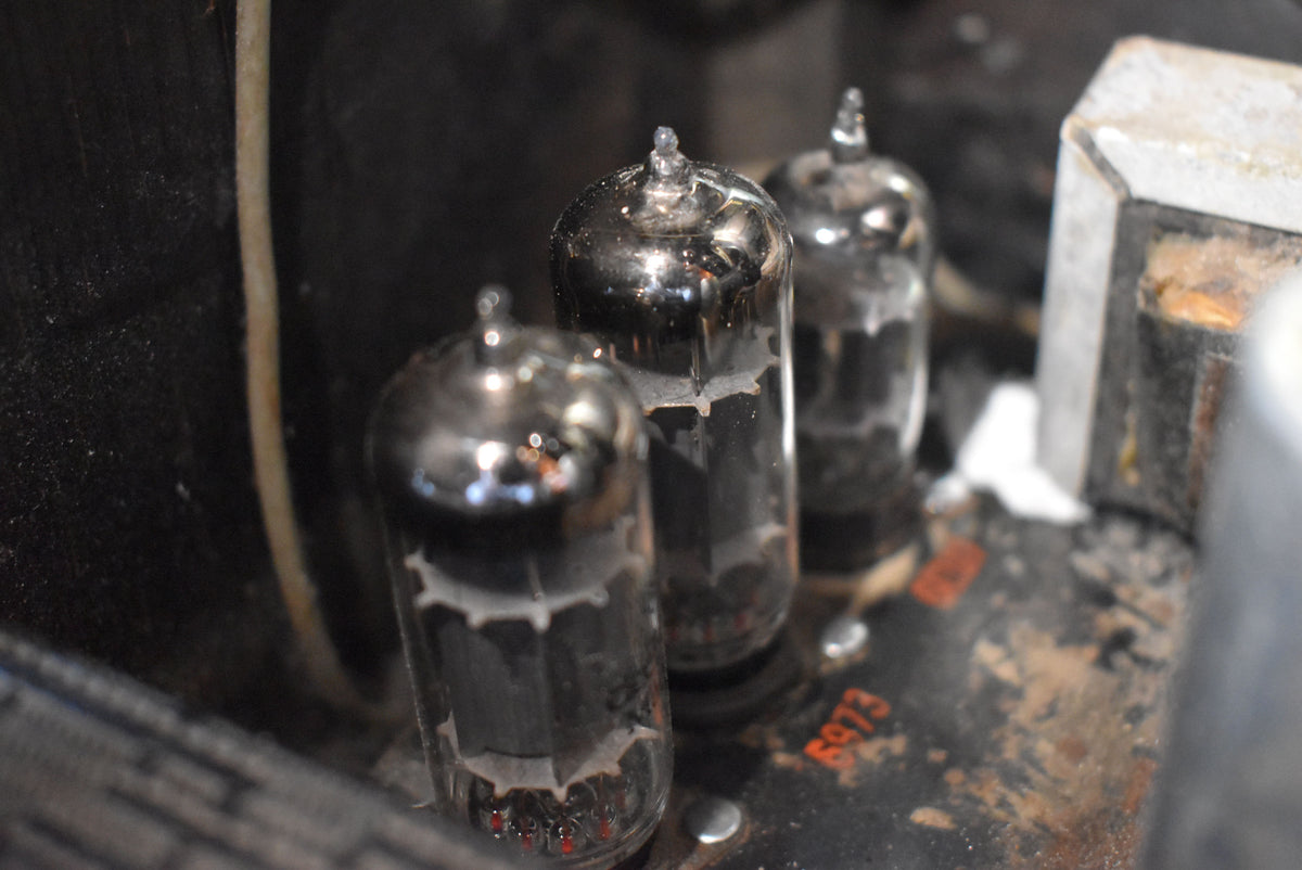 Close-up of three vacuum tubes, showing their glass envelopes and internal elements, mounted on a dusty, worn metal chassis.