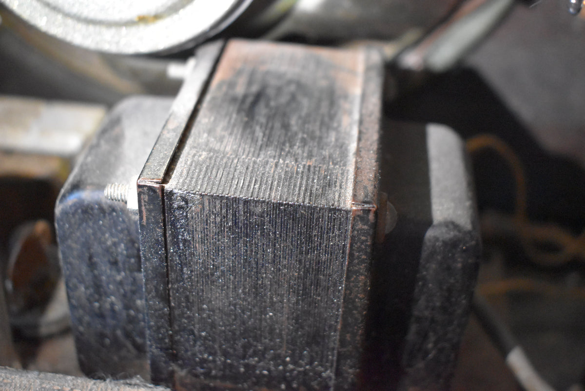 Close-up of a dark, rectangular transformer with visible laminations, secured by a bolt, showing some dust and minor corrosion.