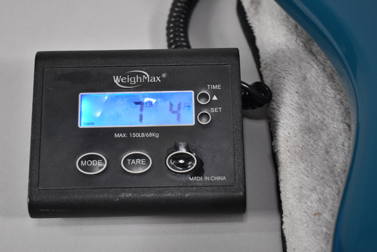 A black digital scale displays "7 LB 4 OZ" with a blue guitar body and white towel visible on the right.