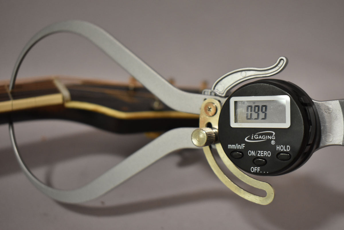 Digital calipers measure the guitar's neck thickness at 0.99 inches, showing the dark wood fretboard and light binding.