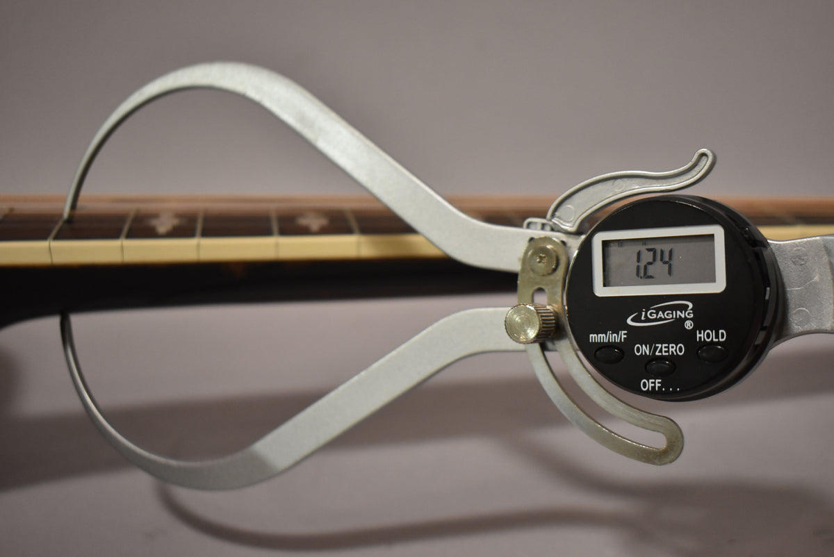 A digital caliper measures the guitar's neck thickness at 1.24 inches, showing the dark fretboard with light block inlays.