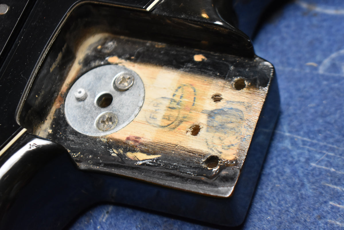 Close-up of the black guitar's neck pocket, showing exposed wood with faint stamps, screw holes, and a metal disc.