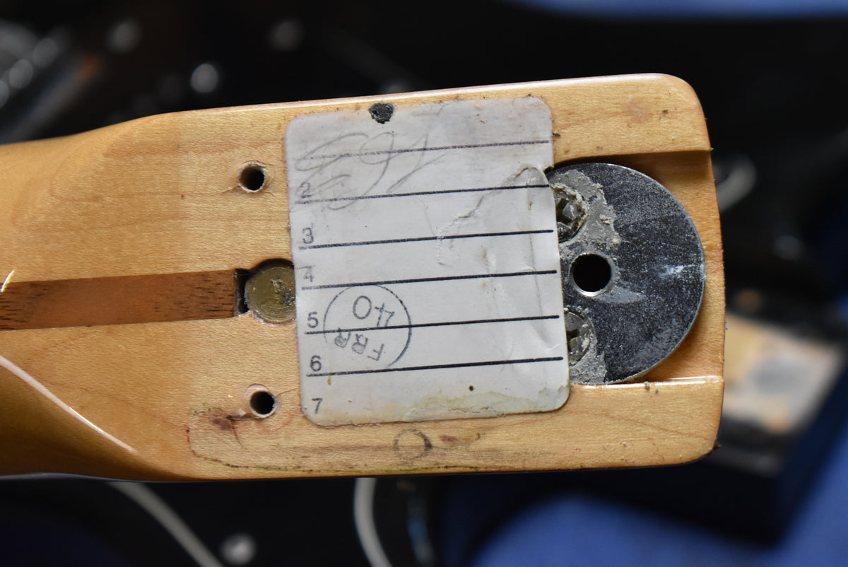 Close-up of the guitar neck heel, showing a light wood finish, a worn paper label, and a metal neck plate with three screws.