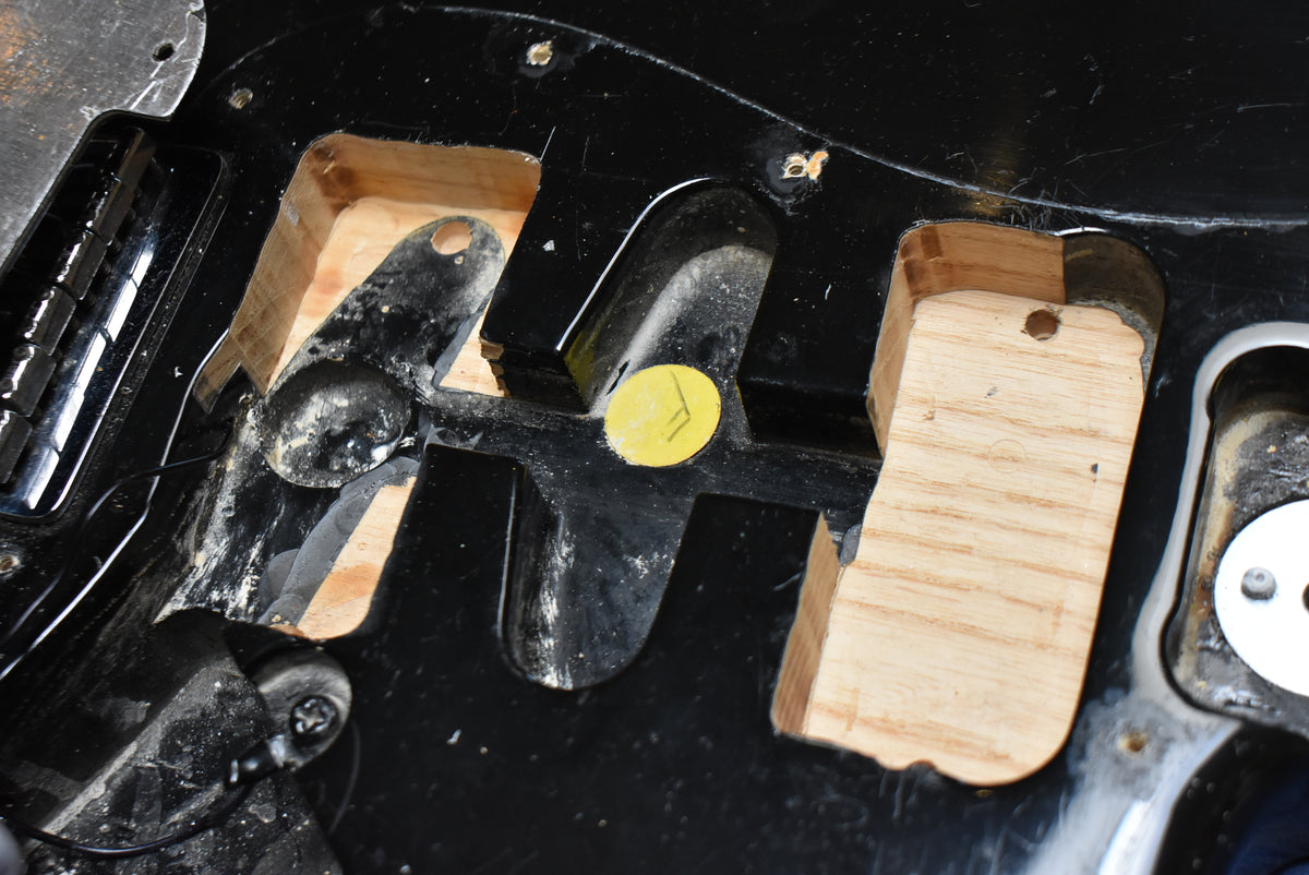 Close-up of the black guitar body's interior, revealing routed wood cavities, a yellow inspection sticker, and some dust and wear.