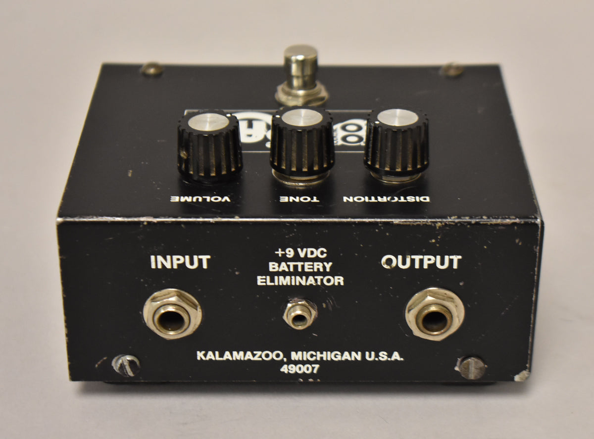 A black distortion pedal is shown from above, with three black ridged knobs, a silver footswitch, and three input jacks.