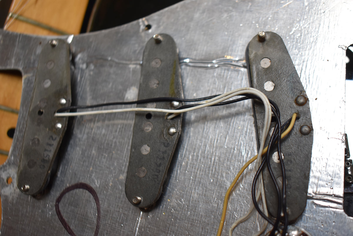 Close-up of three single-coil pickups with black bobbins and exposed pole pieces, wired together over a metallic shielding plate.