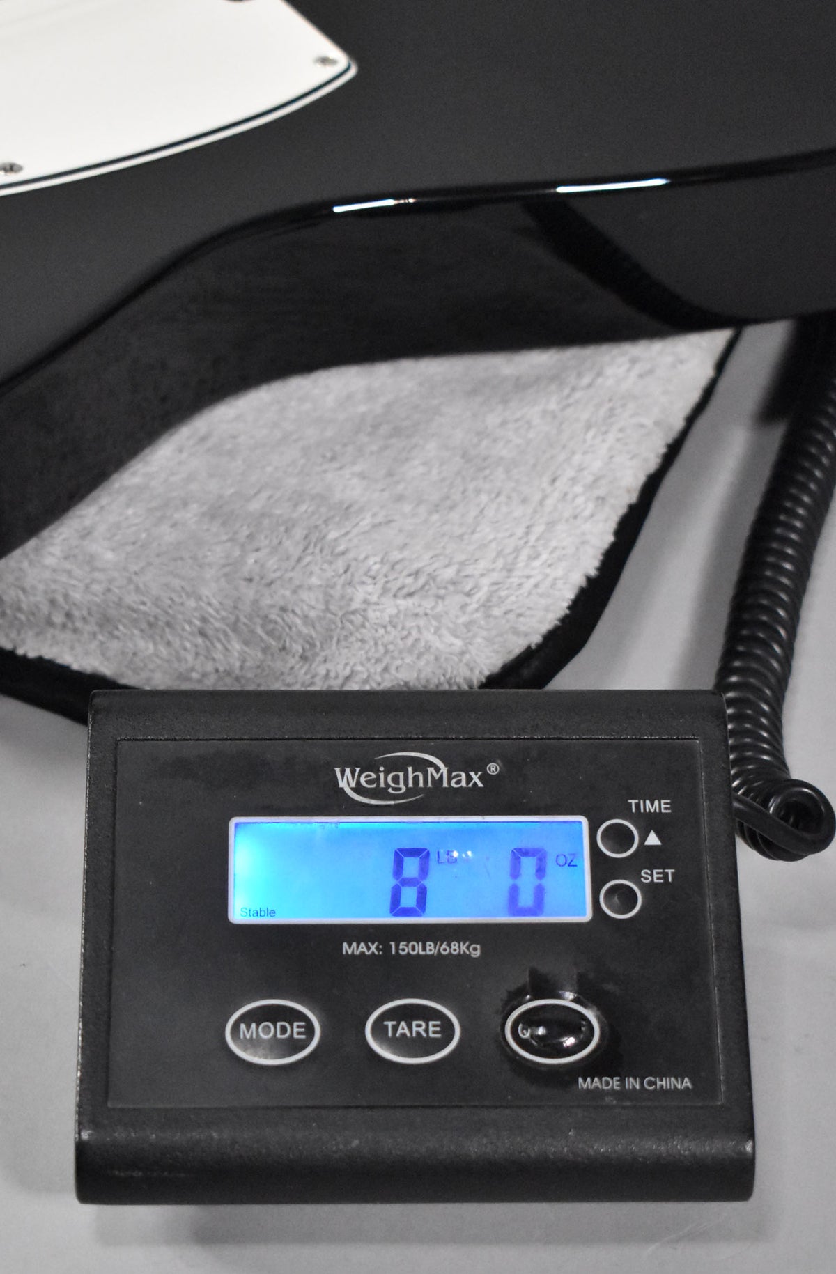 The black Telecaster's lower bout rests on a scale displaying 8 lbs 0 oz, with a white pickguard visible at the top left.