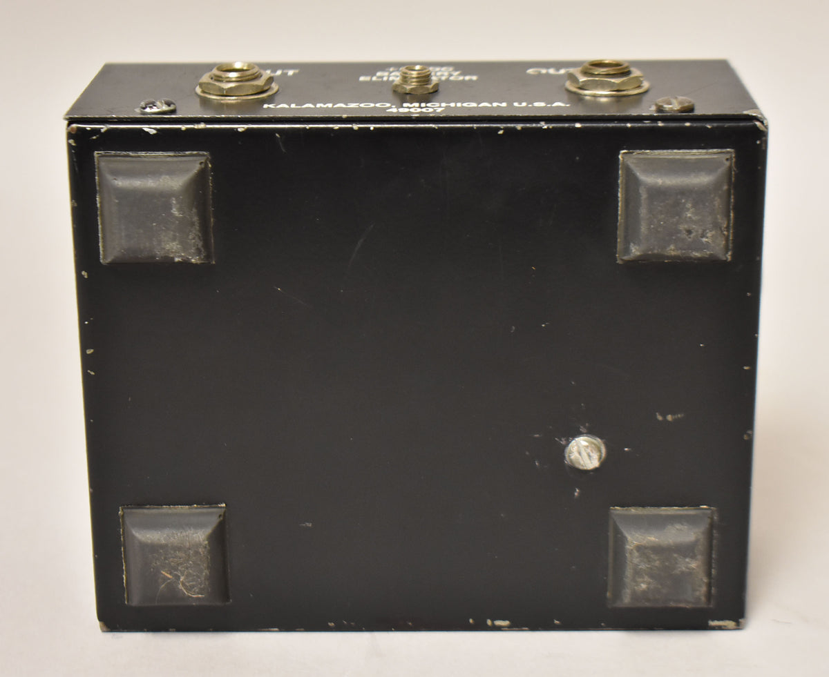 The black metal pedal is viewed from the bottom, showing four worn rubber feet and a silver screw, with input jacks visible on top.