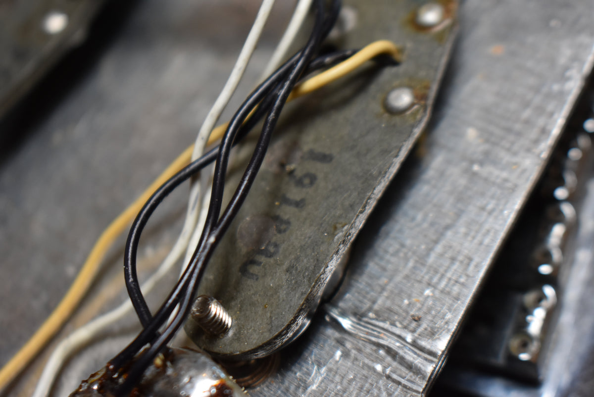 A close-up of a guitar pickup's underside, showing a grey metal plate with black and yellow wires, and a visible screw.