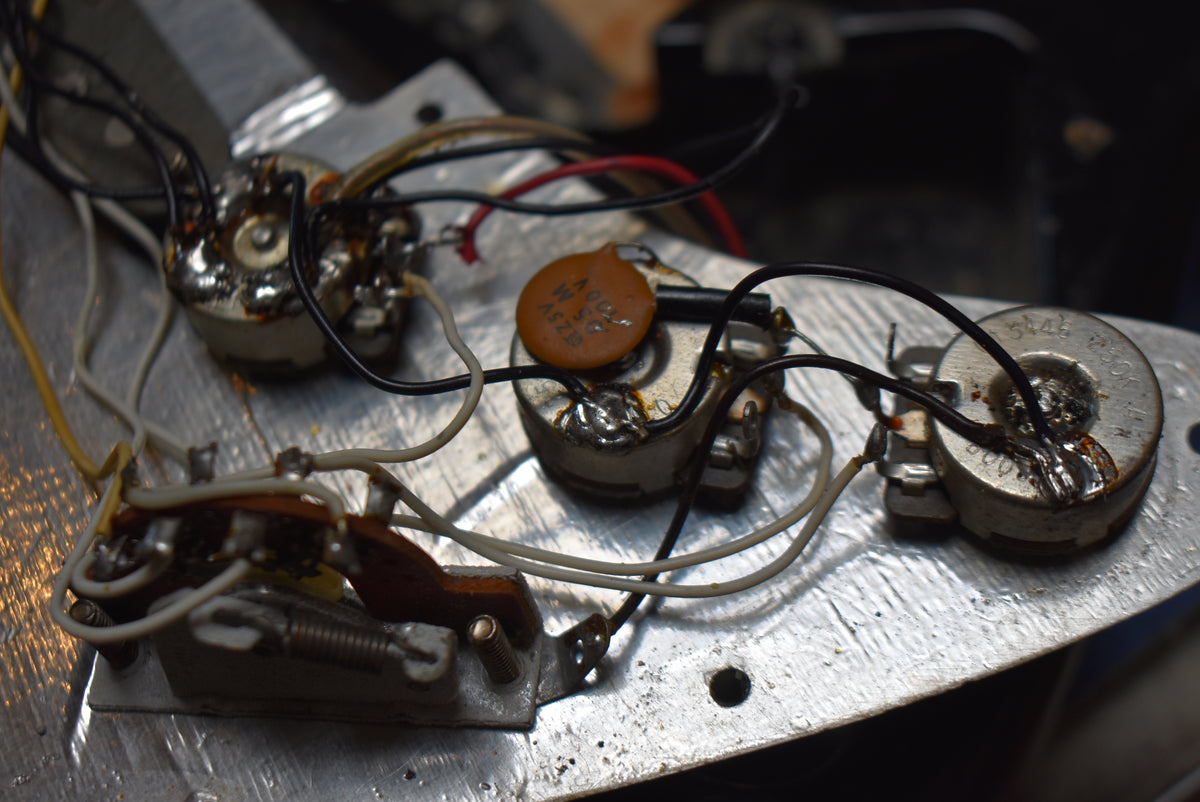 Close-up of the guitar's internal electronics, showing three silver pots, an orange capacitor, and various wires on a metallic plate.