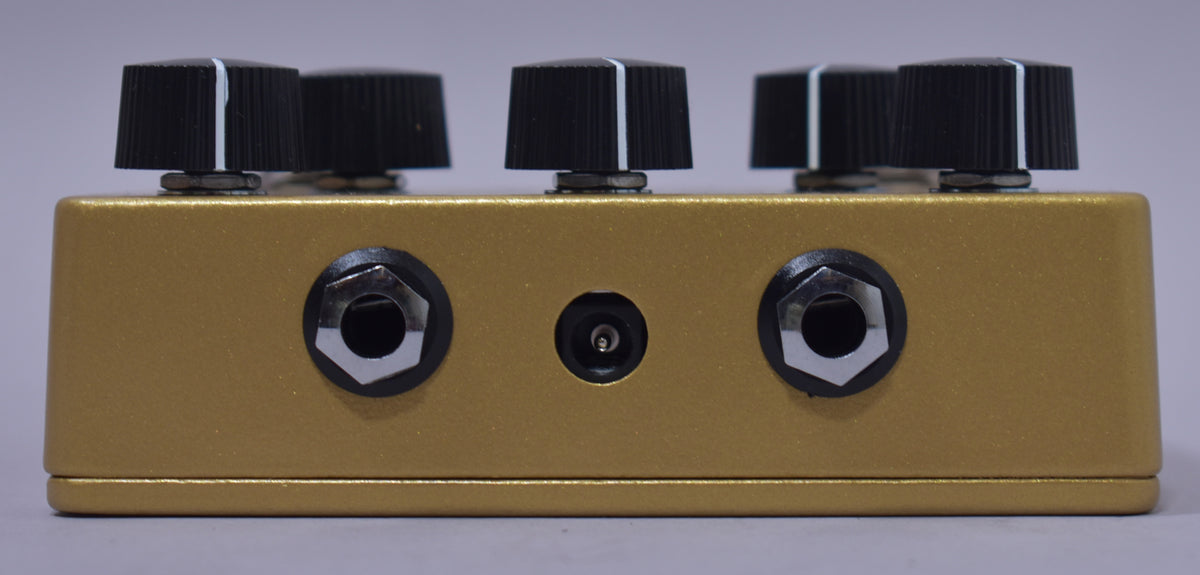 The gold pedal's top edge and rear are visible, showing three black control knobs and two input jacks flanking a power input.