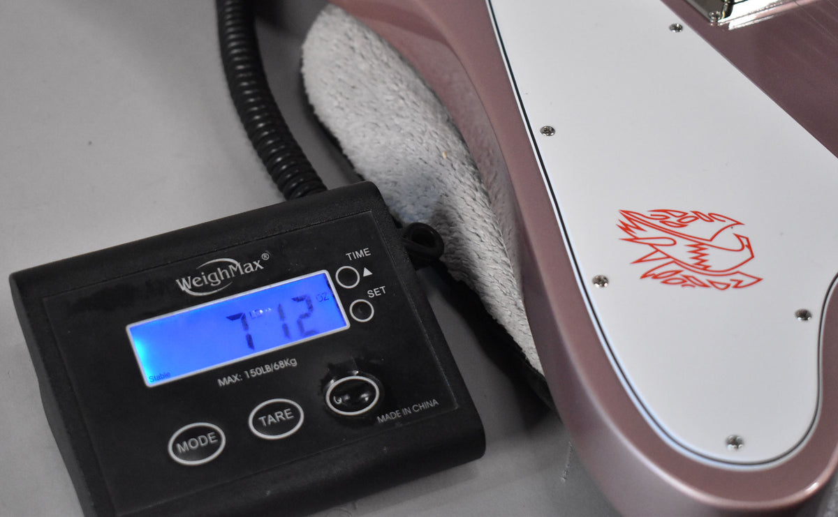 The Firebird's heather poly body and white pickguard with red bird graphic are visible next to a scale displaying "7 LBS 12 OZ".