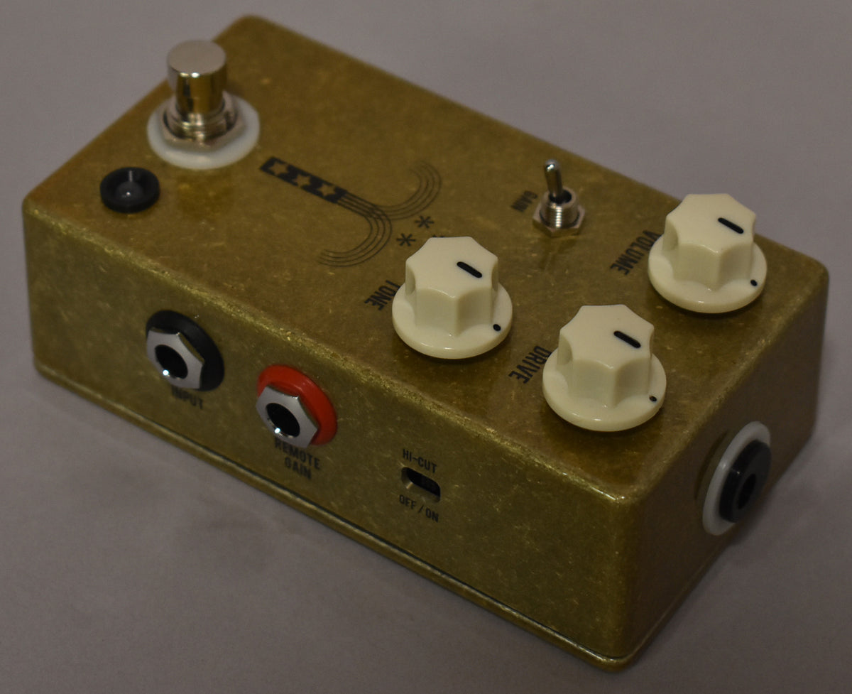 A gold-flecked overdrive pedal, viewed from above and the front, showing three cream knobs, a toggle switch, and input/output jacks.