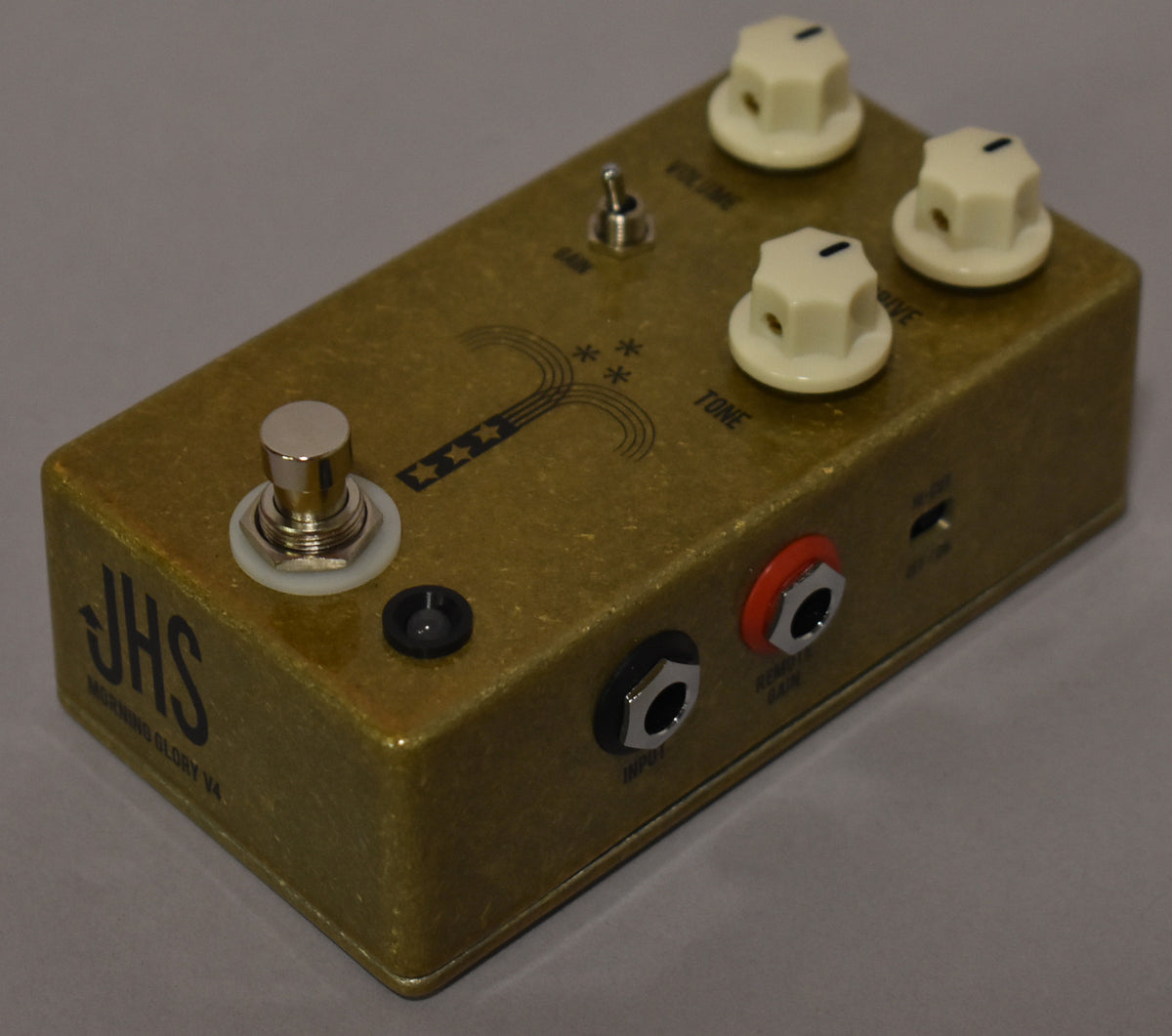 The gold-finished pedal is angled from the front-right, showing three cream knobs, a chrome footswitch, and two input jacks.