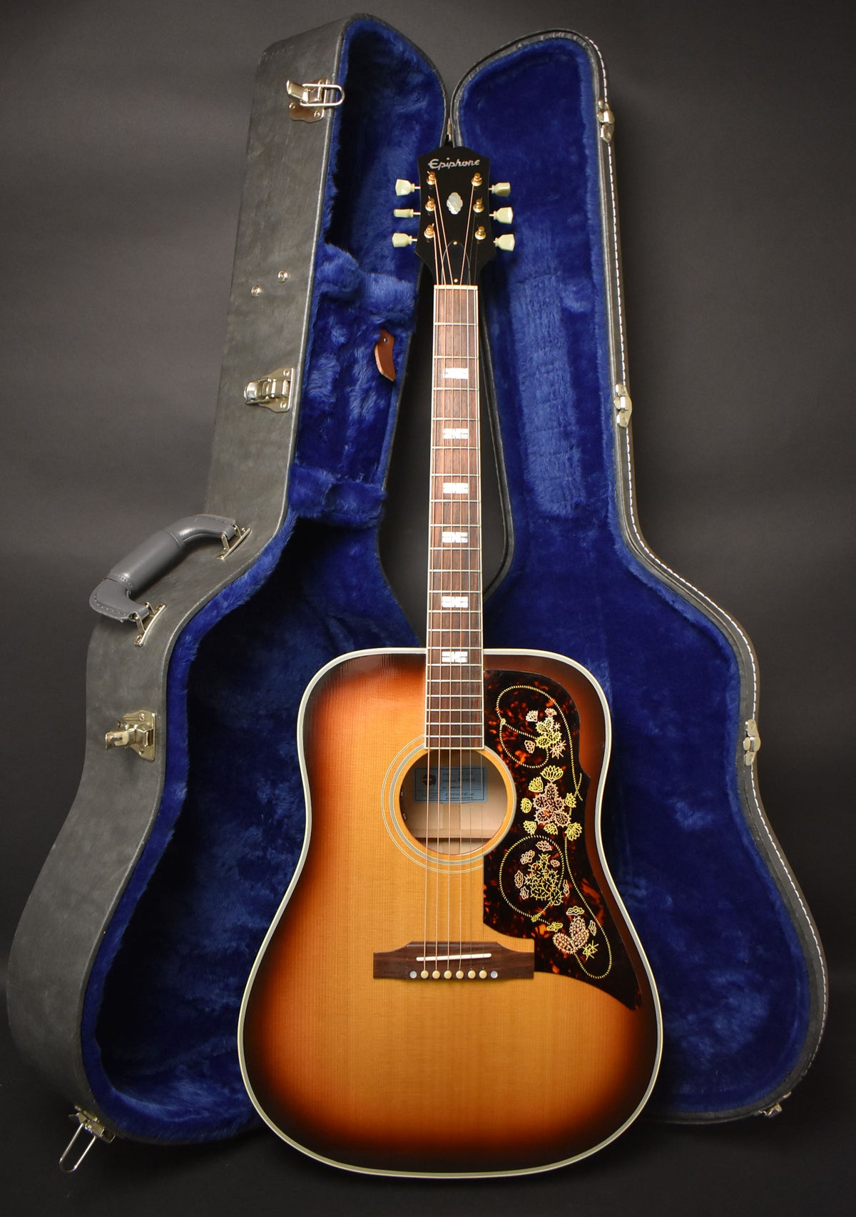 A sunburst acoustic guitar with a decorated pickguard stands upright in front of its open, blue-lined hard case.