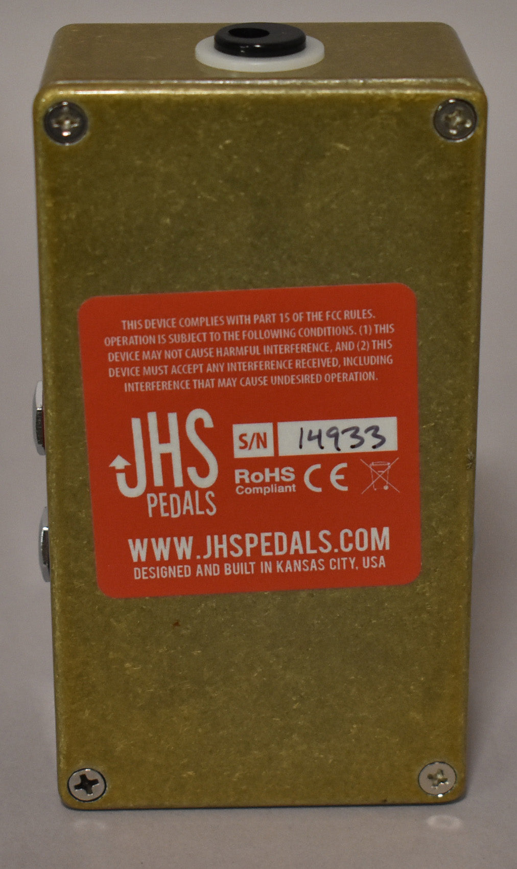 The pedal's bottom is visible, showing a large orange label with text and a serial number, on a textured gold finish.