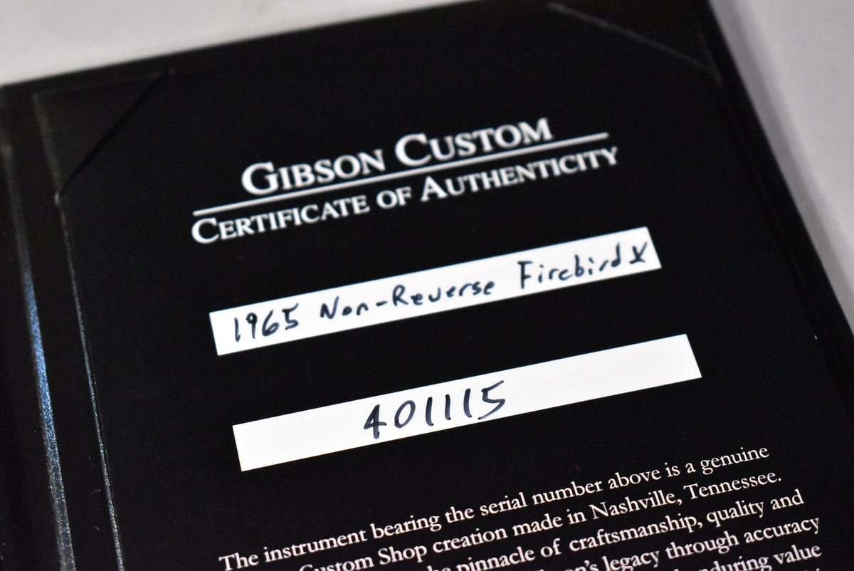 Close-up of a black Gibson Custom Certificate of Authenticity, with "1965 Non-Reverse Firebird" and "401115" handwritten on white labels.